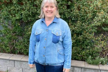 Lady with grey hair wearing light blue denim jacket and dark blue jeans in front of green hedge and grey stone wall.