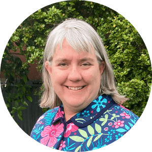 Carmen Grantham - grey haired lady wearing floral jacket with green bushes in the background.